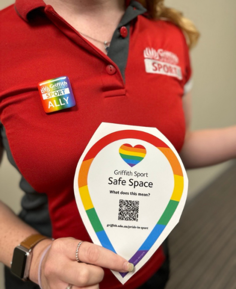 Griffith Sport Staff in Uniform. Wearing Griffith Sport Ally Badge and holding a Griffith Sport Safe Space Sticker. 