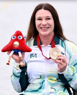 An image of a happy female wearing the Australian Paralympics uniform holding up a bronze medal and Paris 2024 mascot plush toy