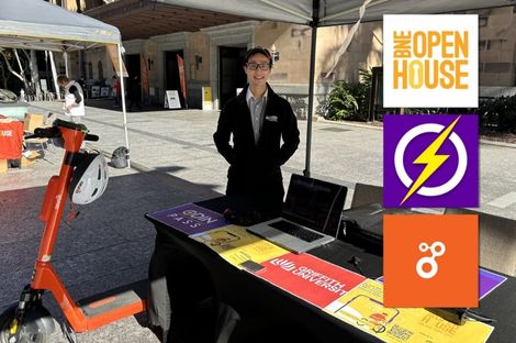 a man standing in a market booth with flyers, laptop and e-scooter